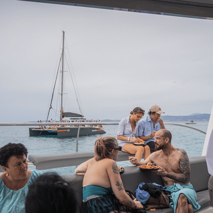 Group enjoying a meal on the deck of an Attraction Catamarans catamaran, with views of the Mallorca coast and other vessels.